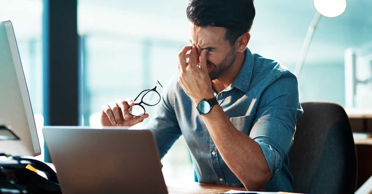 A man wearing a button-down shirt sits at a desk with a laptop and a monitor. He takes off his glasses and rubs his eyes.