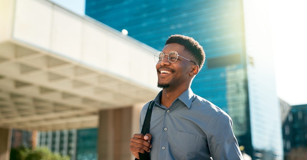 A man smiles and walks with a bag on his shoulder. Tall office buildings and a ray of sunlight are in the background.